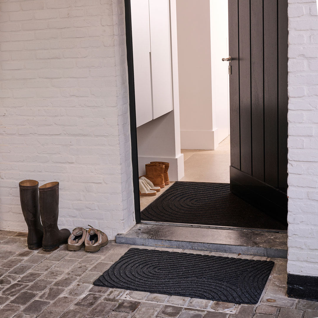 Black doormat in front of a house entrance with shoes and boots nearby.