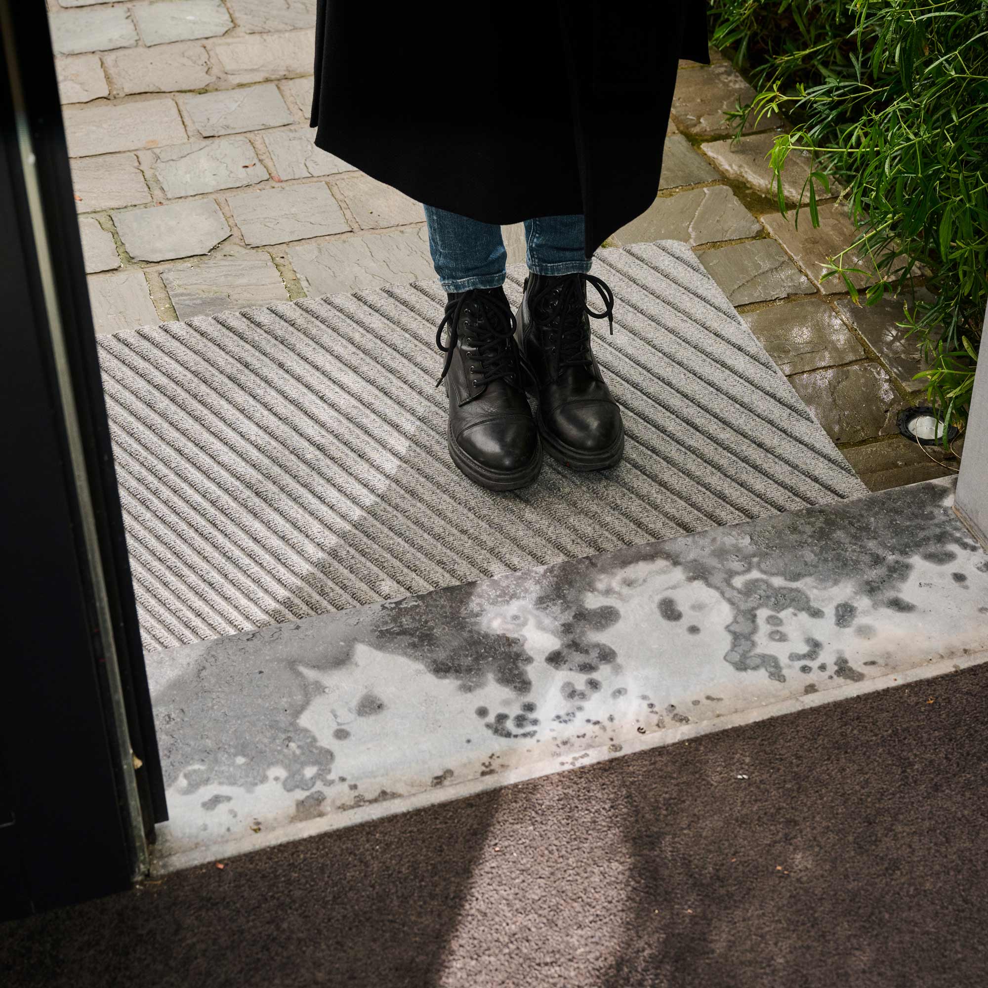Person wearing black boots stepping onto a doormat with a wet ground outside.