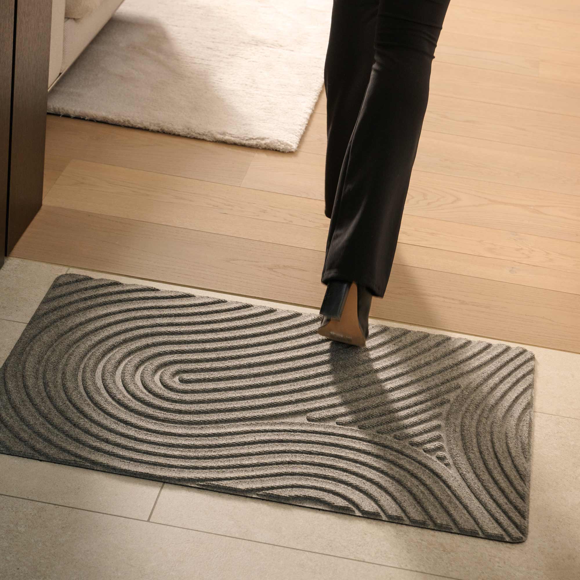 Person stepping onto a textured doormat with wavy pattern on a wooden floor.