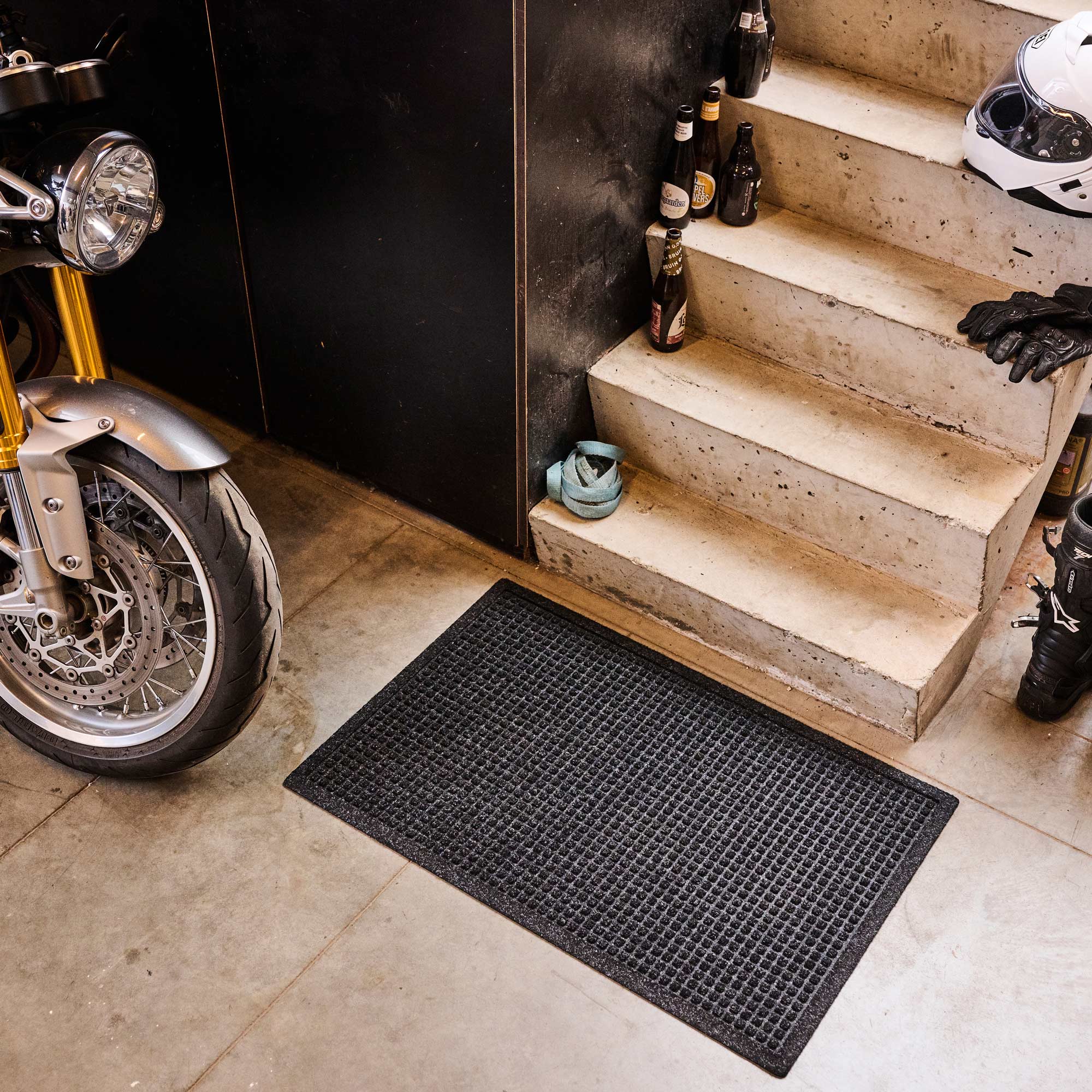 Black doormat on a concrete floor with a motorcycle and stairs in the background.