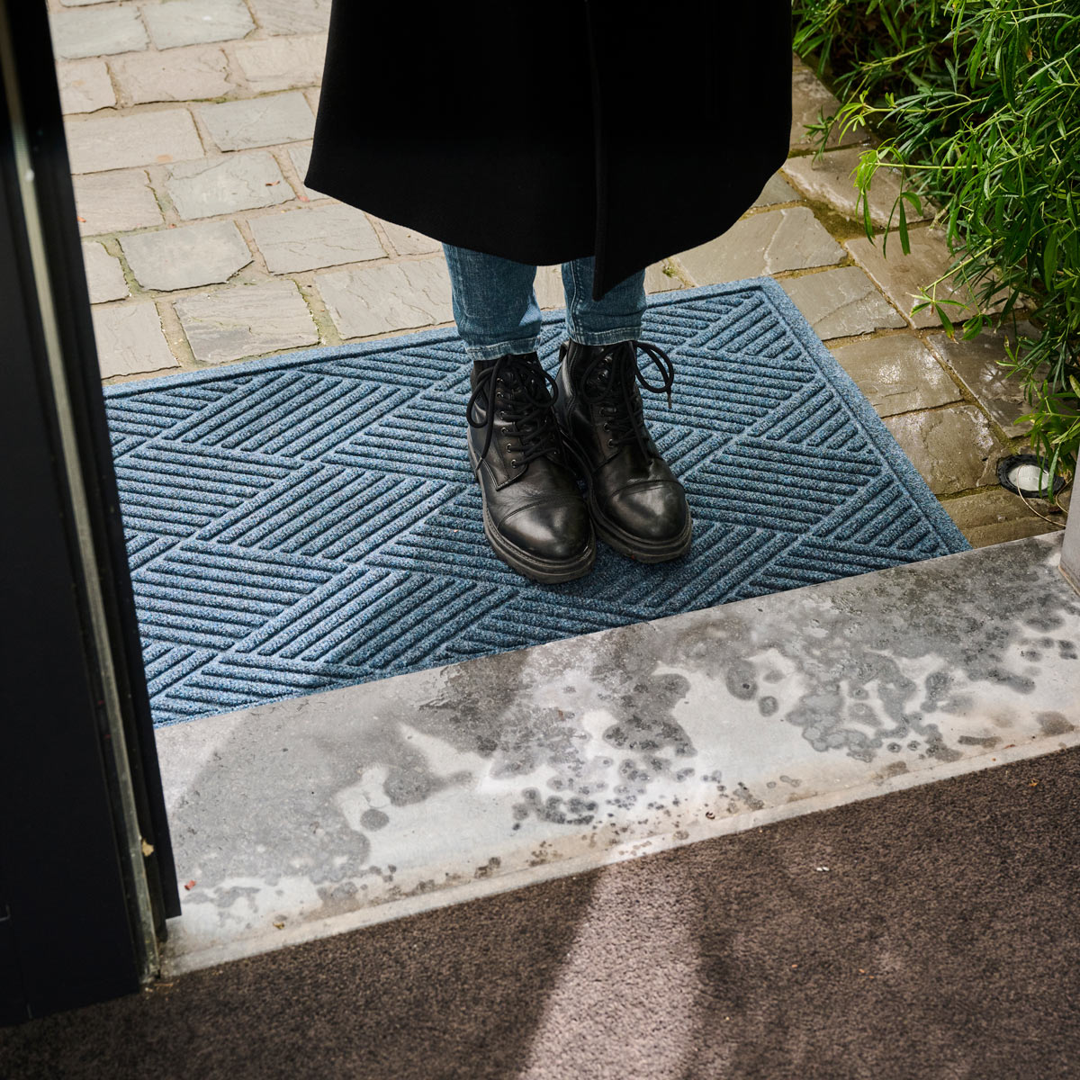 Person wearing black boots stepping onto a blue doormat from a door.
