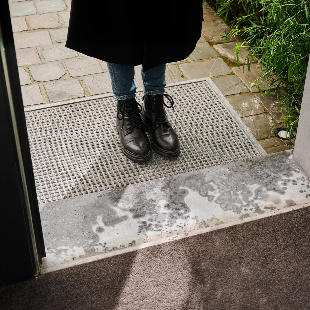 Person wearing dark boots and a coat standing on a textured mat outside a building.